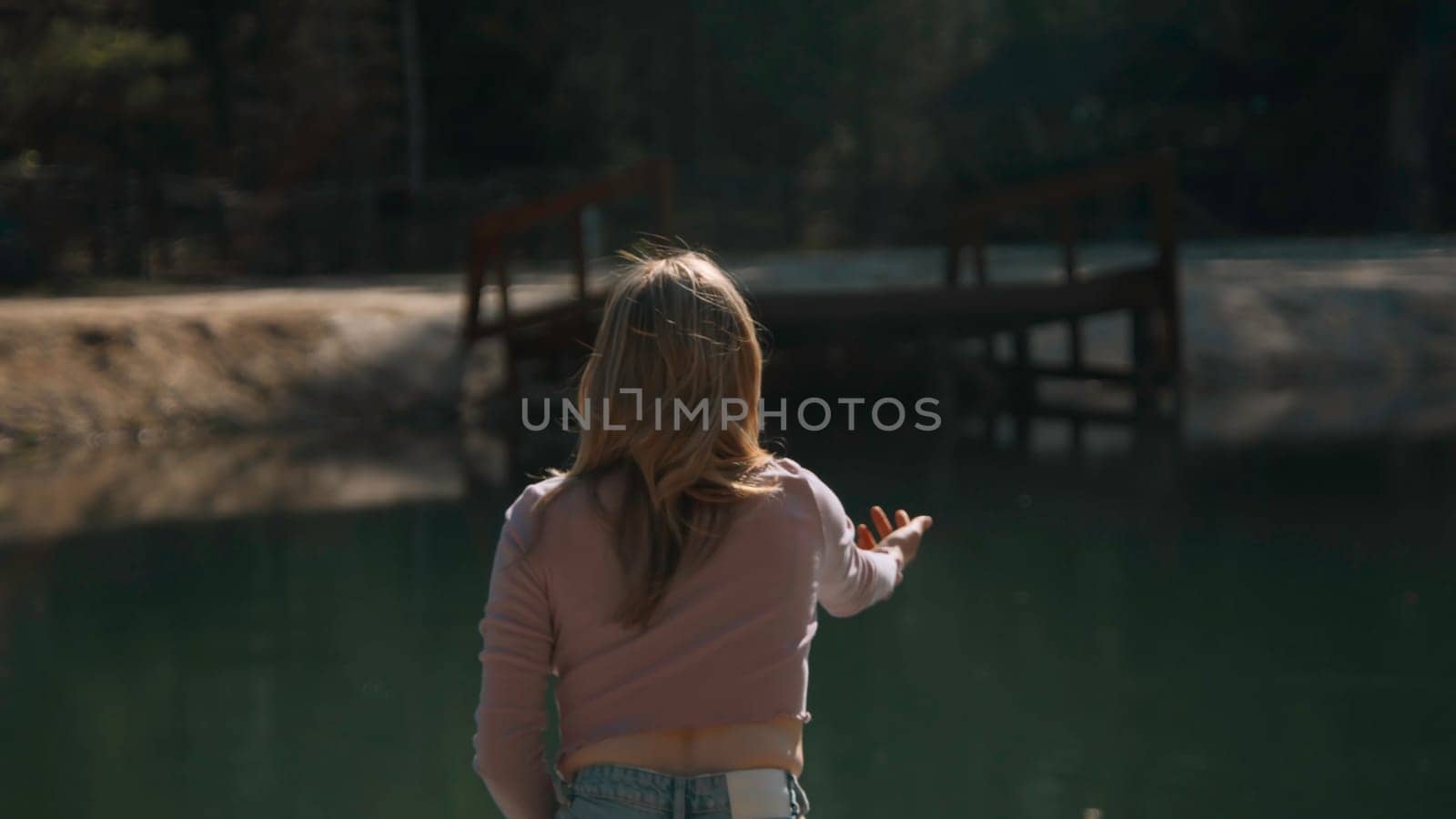 Rear view of young woman throwing rocks into water. Stock footage. Attractive young woman sits on pier and throws stones. Woman throws stones on shore of lake on sunny summer day.