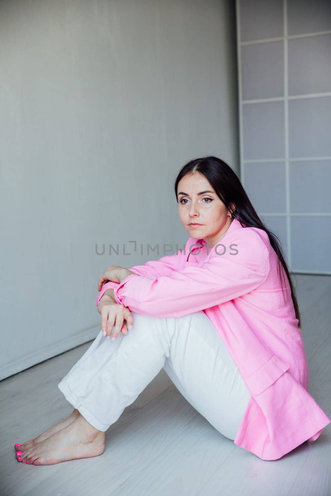 Beautiful fashionable brunette woman sitting on the floor in pink jacket and white pants