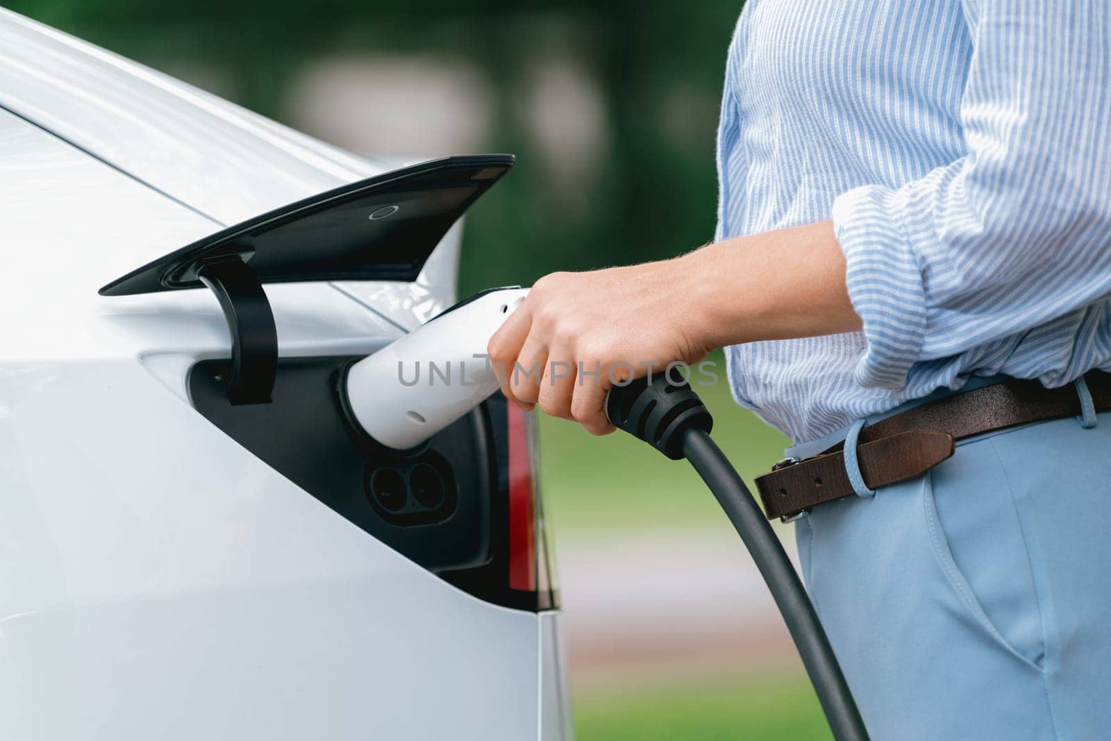 Young woman recharge EV electric vehicle's battery from EV charging station in outdoor green city park scenic. Eco friendly urban transport and commute with eco friendly EV car travel. Exalt