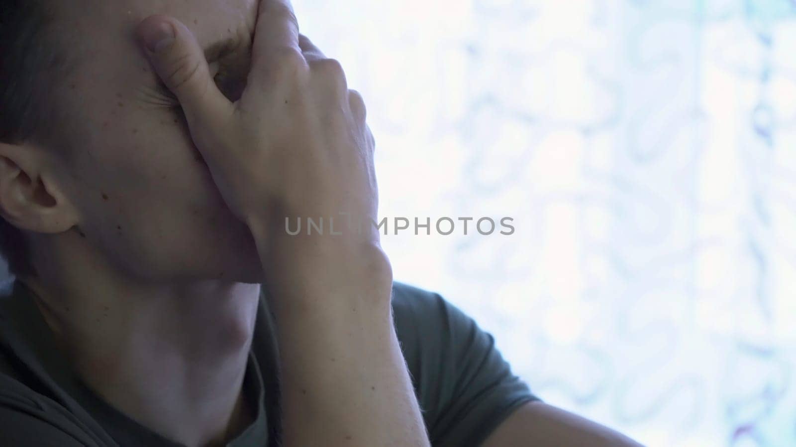 Sad cheerless man, sitting in the room and covering his face. Close-up