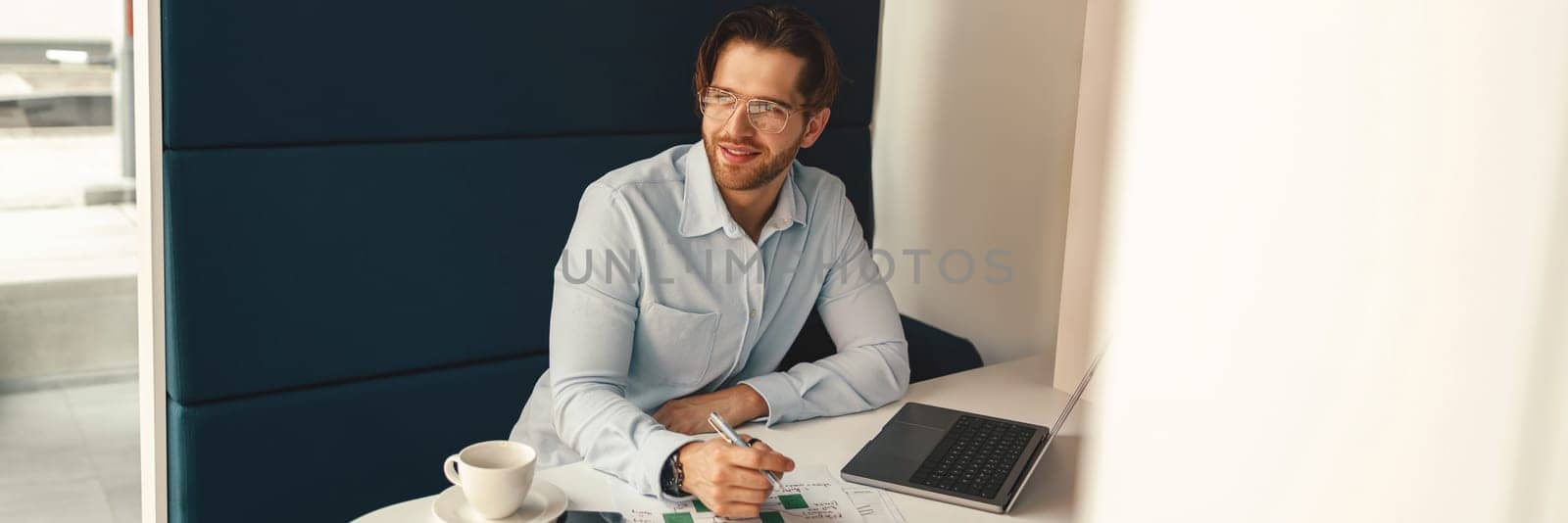 Handsome smiling male freelancer making notes during working on laptop sitiing desk in coworking
