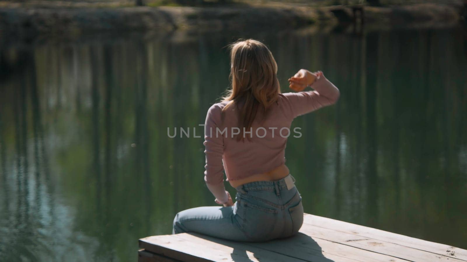 Beautiful young woman throws bread into lake. Stock footage. Attractive woman sits on pier and throws stones into lake. Young woman throws stones into lake on sunny summer day.