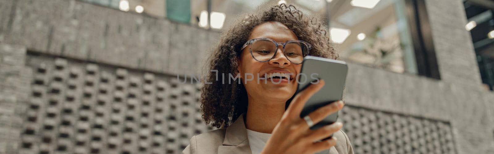 Smiling businesswoman is looking on phone while standing on office background