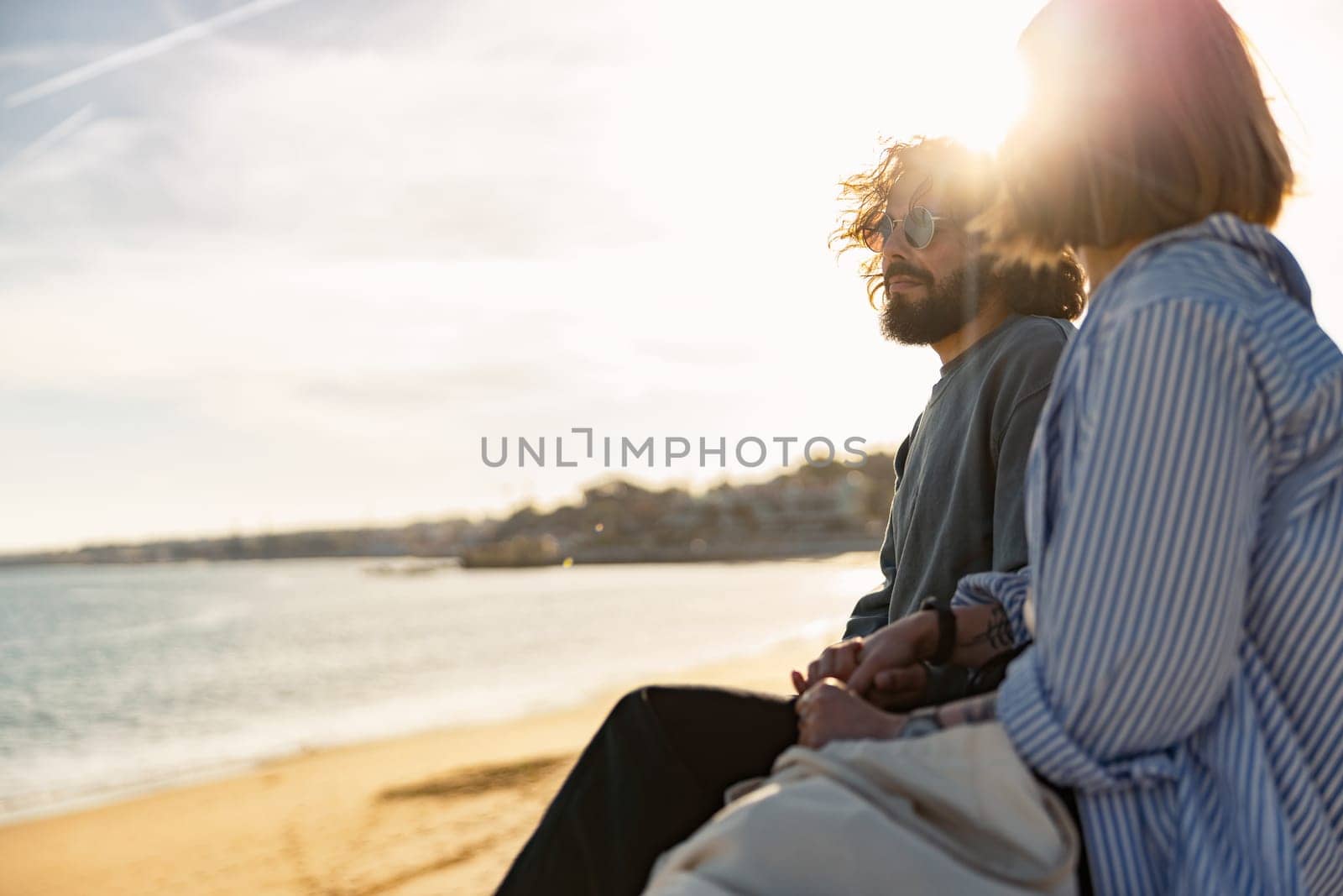 Beautiful romantic couple sitting at the beach wearing casual clothes and looking at the ocean by Yaroslav_astakhov