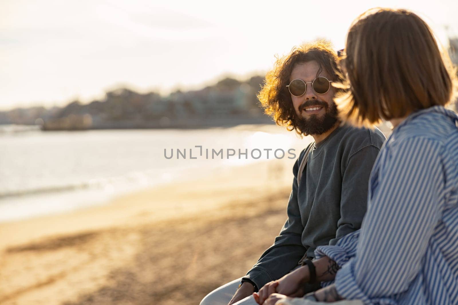 Smiling romantic couple sitting at the beach wearing casual clothes and looking each other by Yaroslav_astakhov