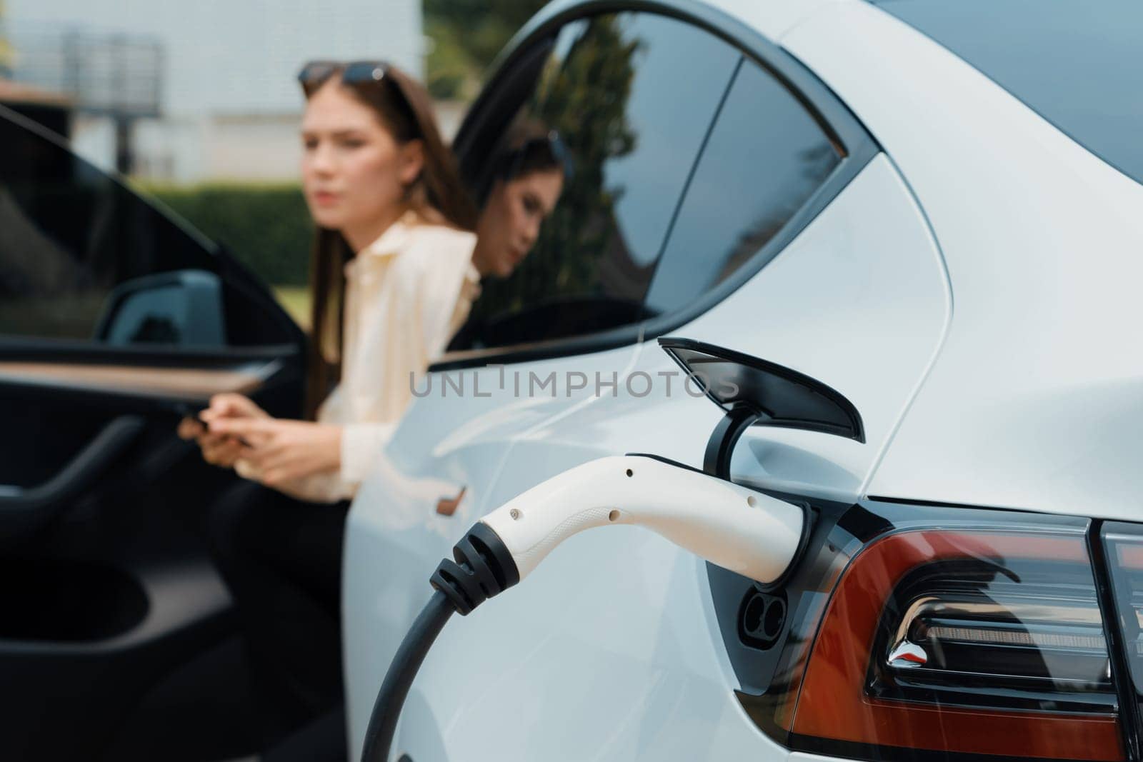 Young woman use smartphone to pay for electricity at public EV car charging station green city park. Modern environmental and sustainable urban lifestyle with EV vehicle. Expedient