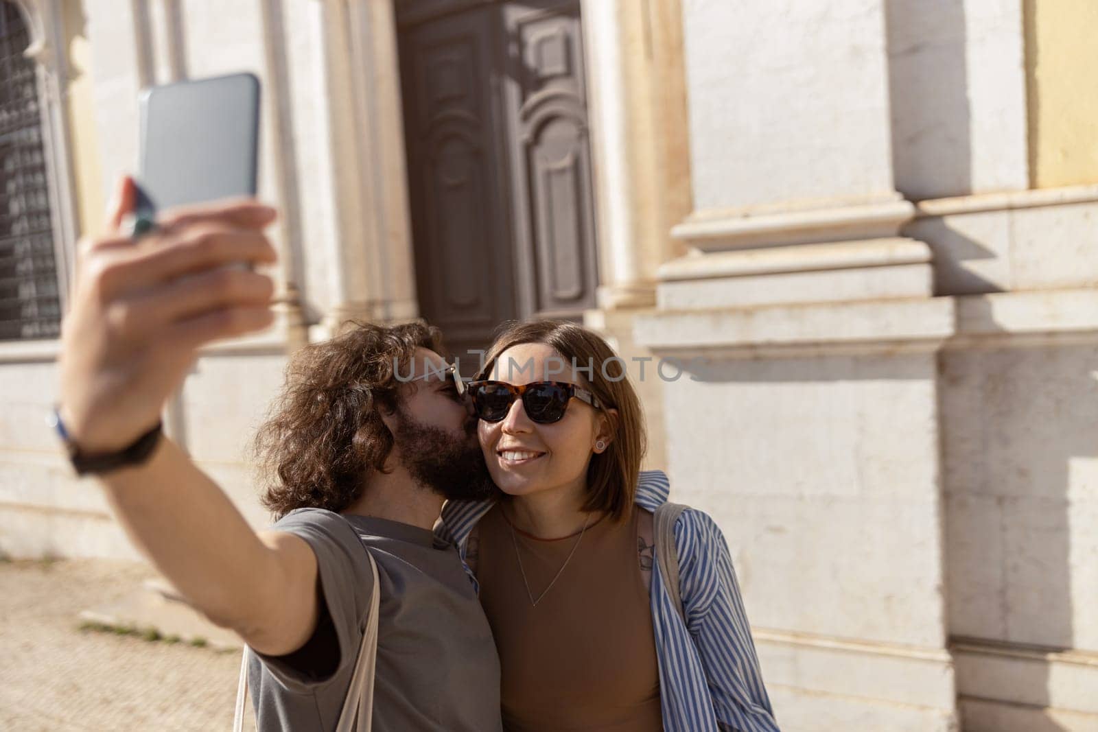 Happy tourist couple walking in city street, kissing and making selfie on phone by Yaroslav_astakhov