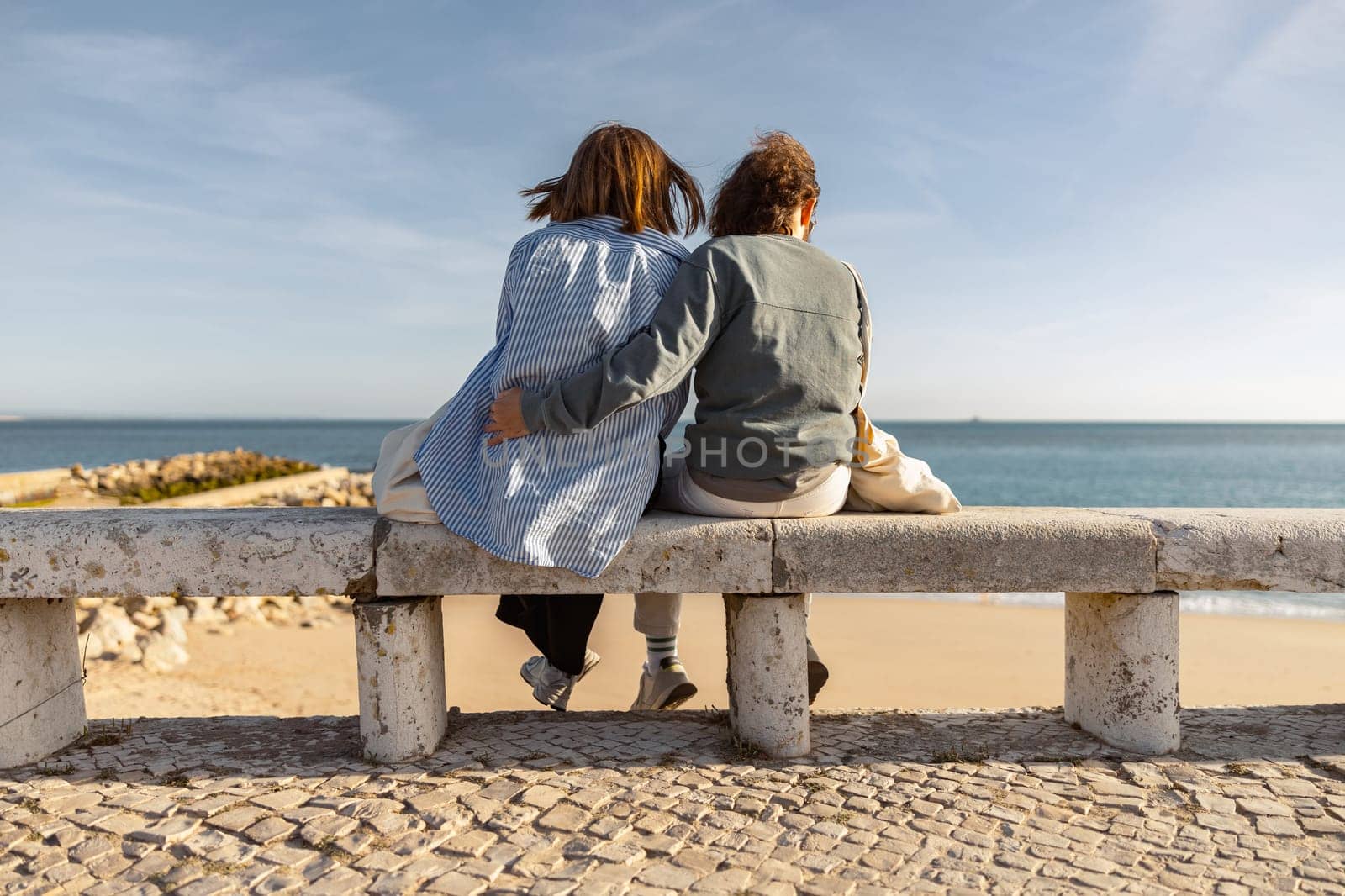 Rear view of couple hugs while sitting on bench and enjoys beautiful ocean view. High quality photo