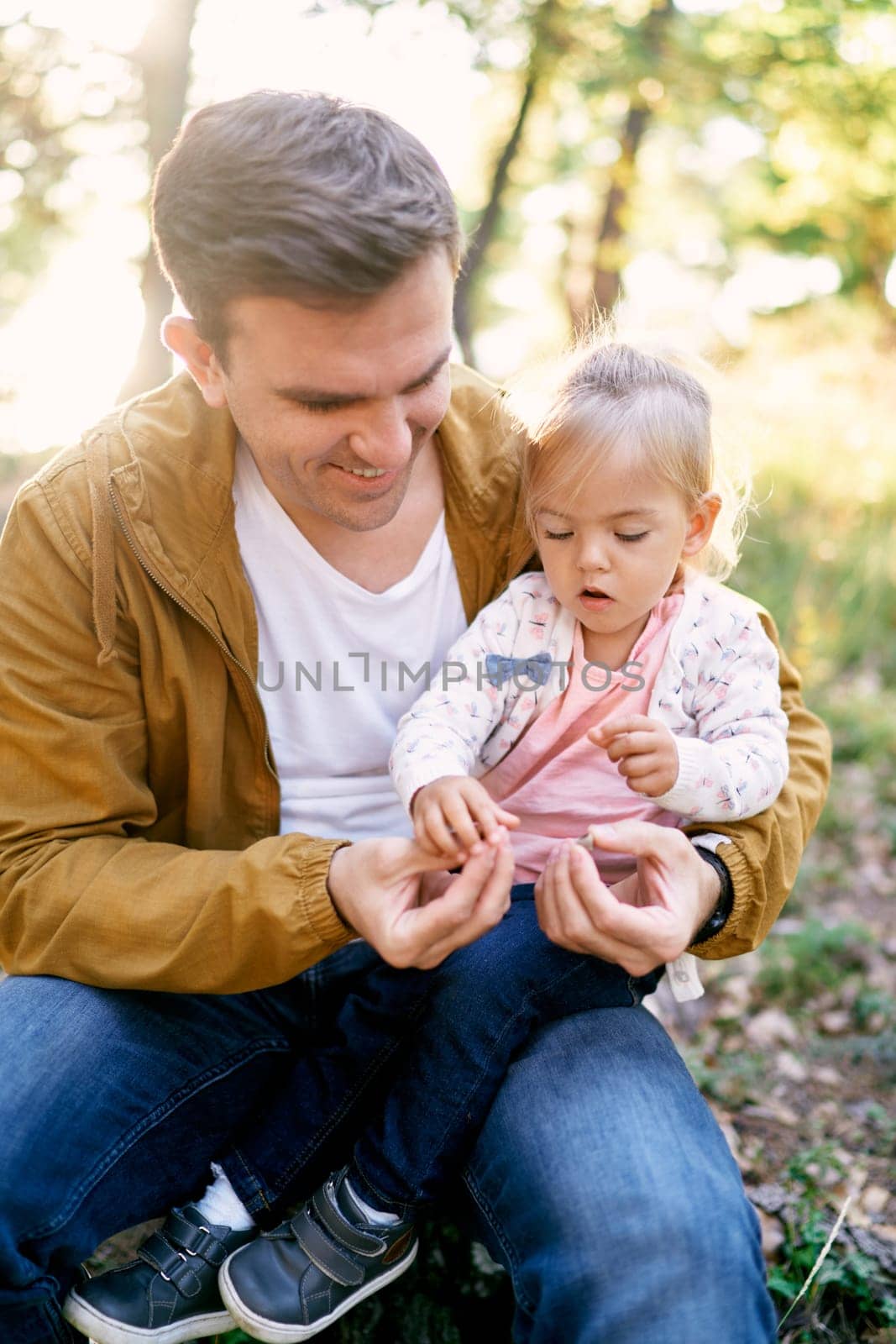 Little girl sits on her father lap in the forest and takes with stones from his hand. High quality photo