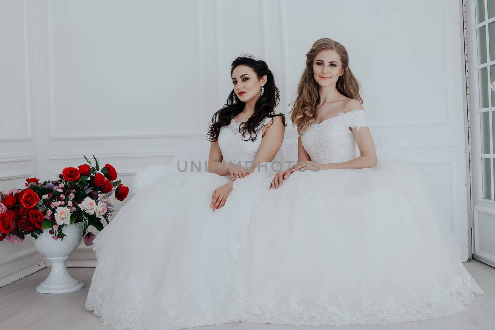 two brides in wedding dresses sit on a white couch in the room