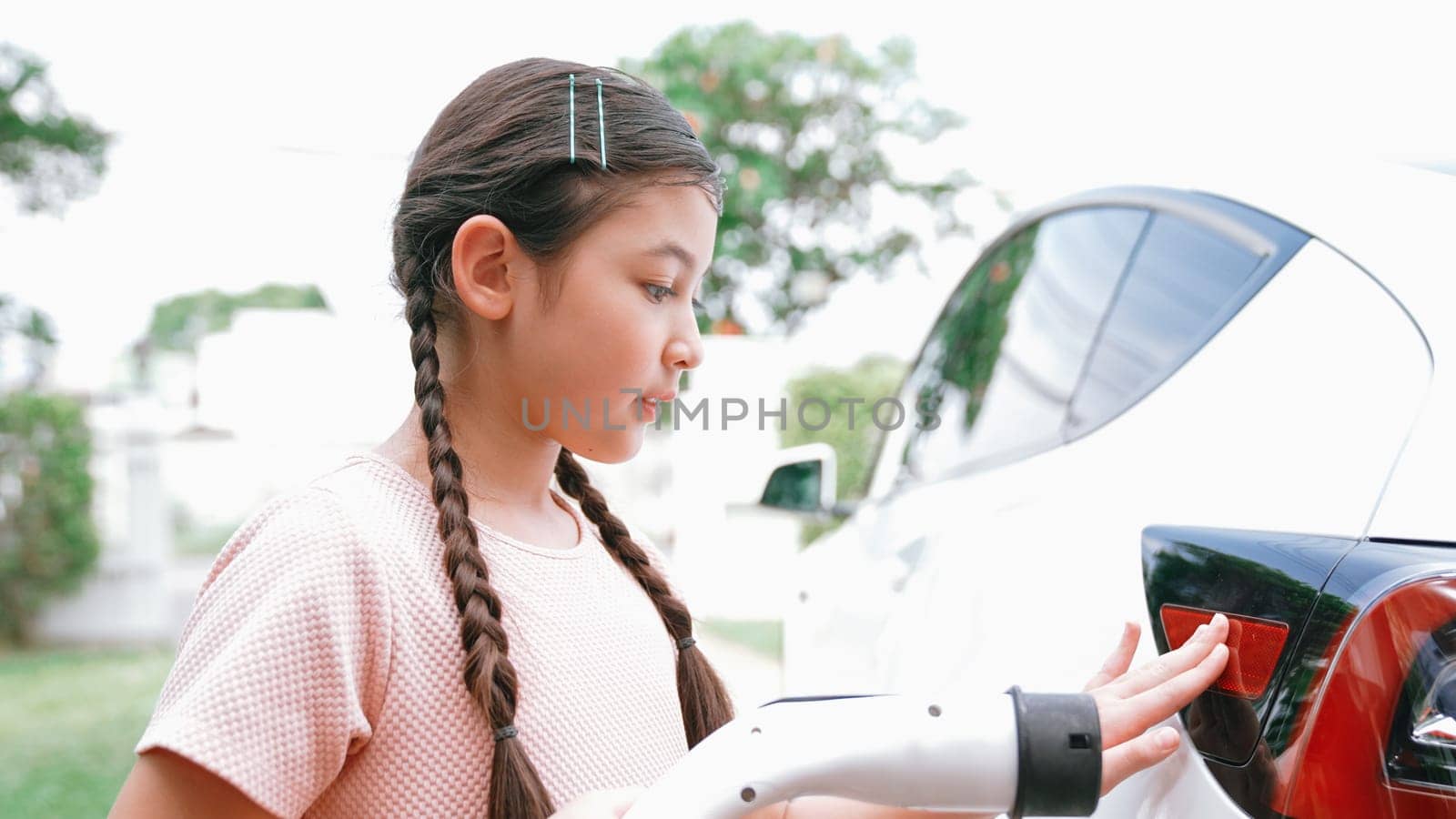 Happy little young girl learn about eco-friendly and energy sustainability as she recharge electric vehicle from home EV charging station. EV car and sustainable future generation concept. Synchronos