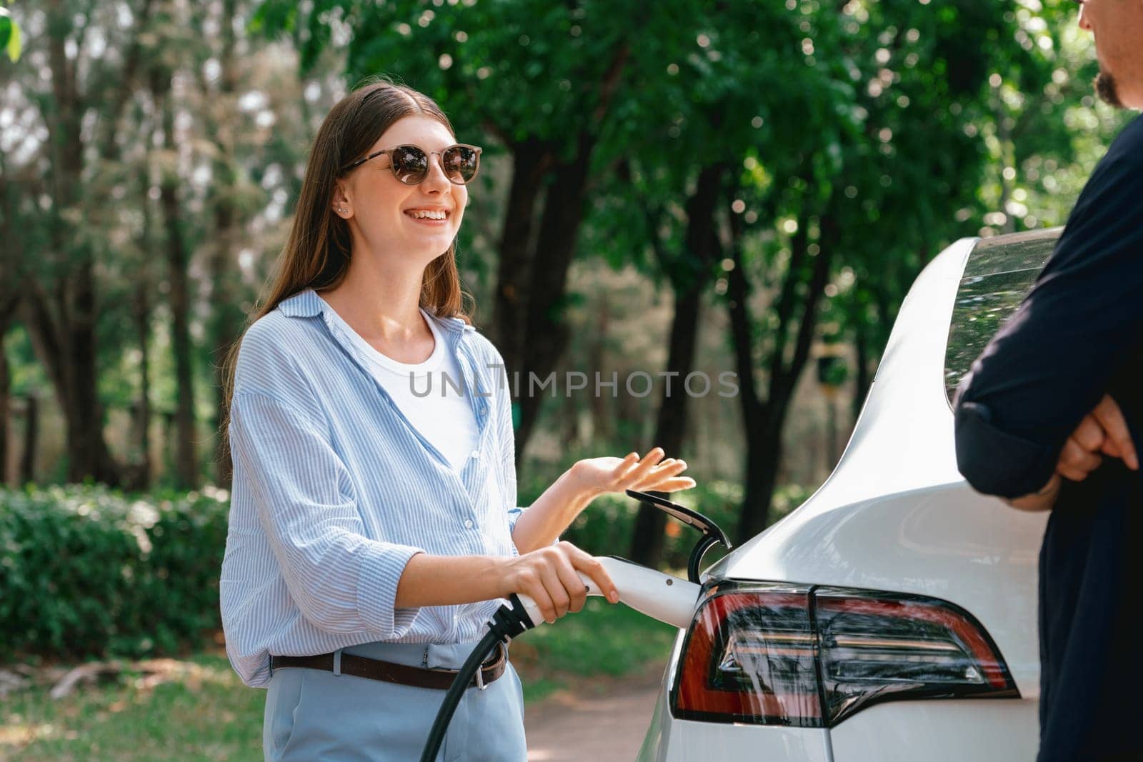 Lovely young couple wearing sun glasses recharging battery for electric car during road trip travel EV car in natural forest or national park. Eco friendly travel during vacation and holiday. Exalt