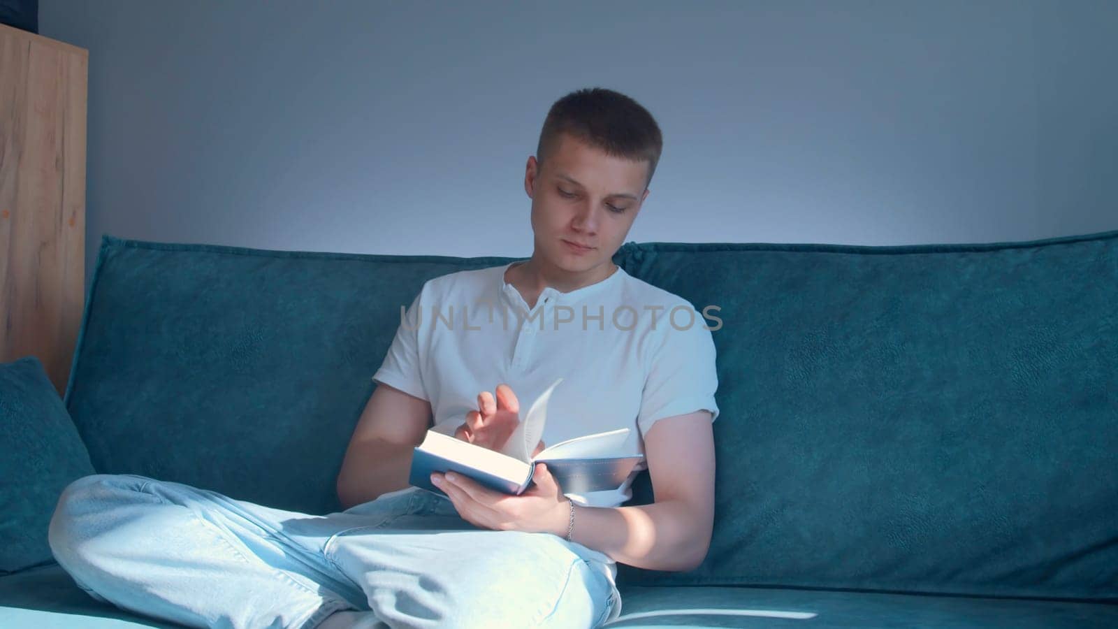 Young man is seriously reading book at home. Media. Handsome man is reading book sitting on couch at home. Young man is reading book and engaged in self-development.