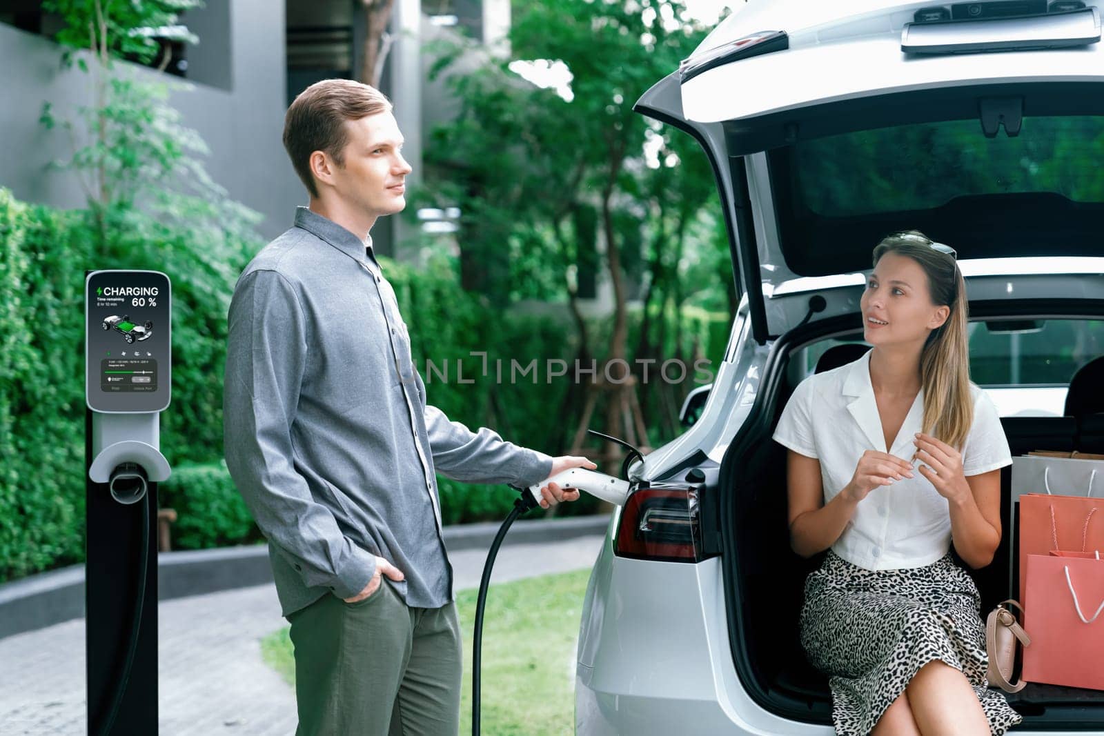 Young couple travel with EV electric car charging in green sustainable city outdoor garden in summer shows urban sustainability lifestyle by green clean rechargeable energy of electric vehicle innards