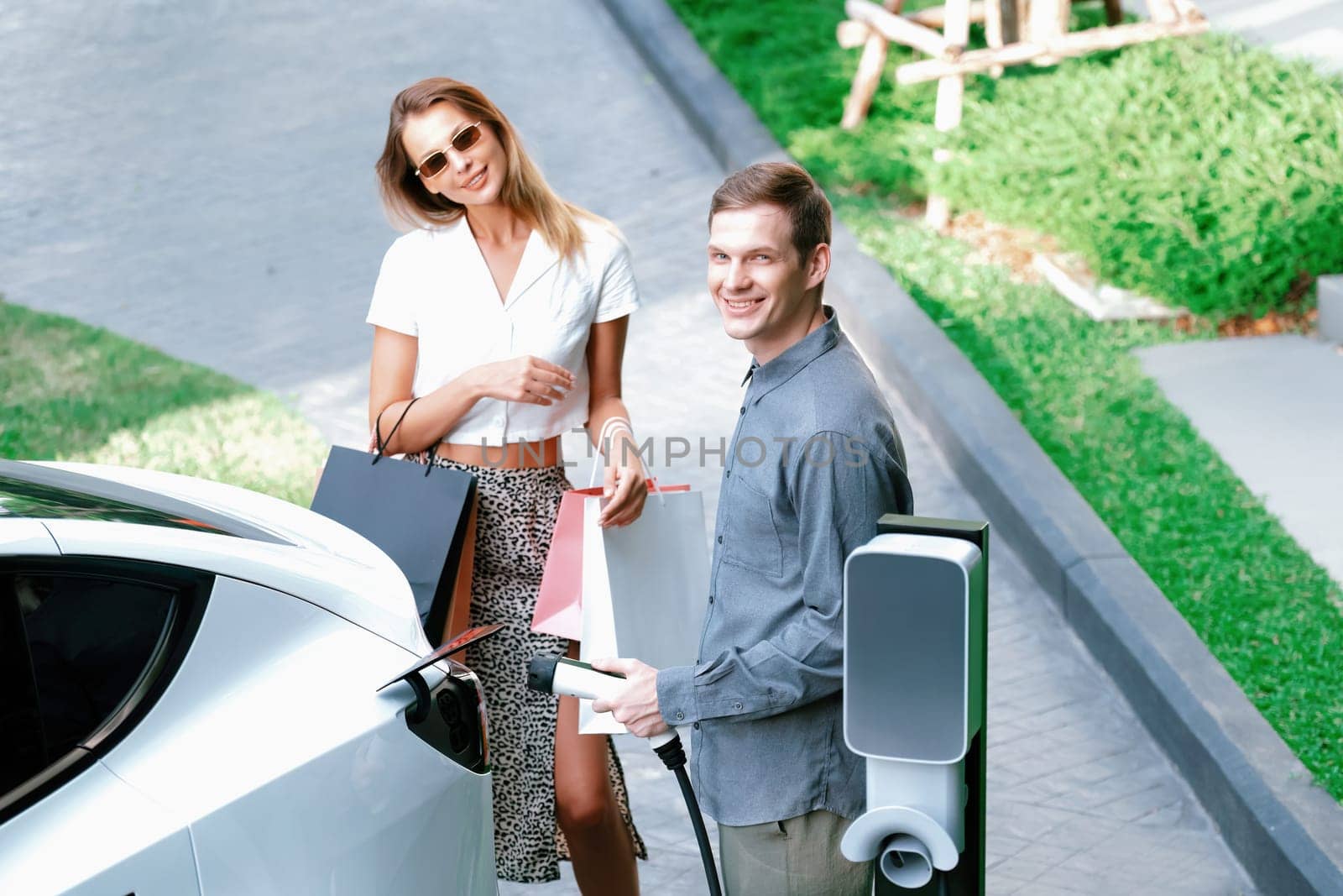 Young couple travel with EV electric car charging in green sustainable city outdoor garden in summer shows urban sustainability lifestyle by green clean rechargeable energy of electric vehicle innards