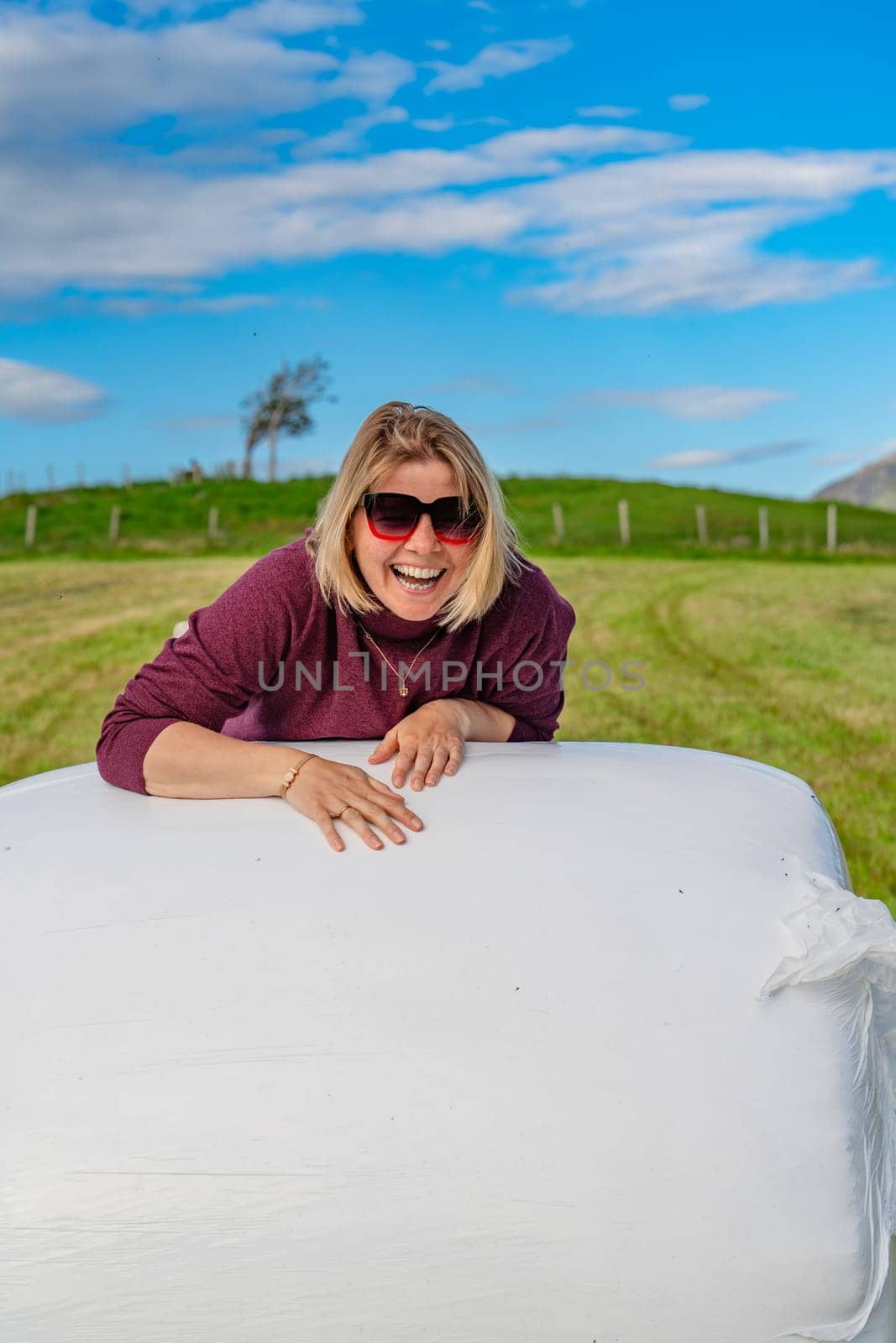 A 40-year-old woman basking in the tranquil aura, casually seated on a densely packed heap of hay in the midst of a charming farm field landscape, revealing a sense of peace and natural elegance.