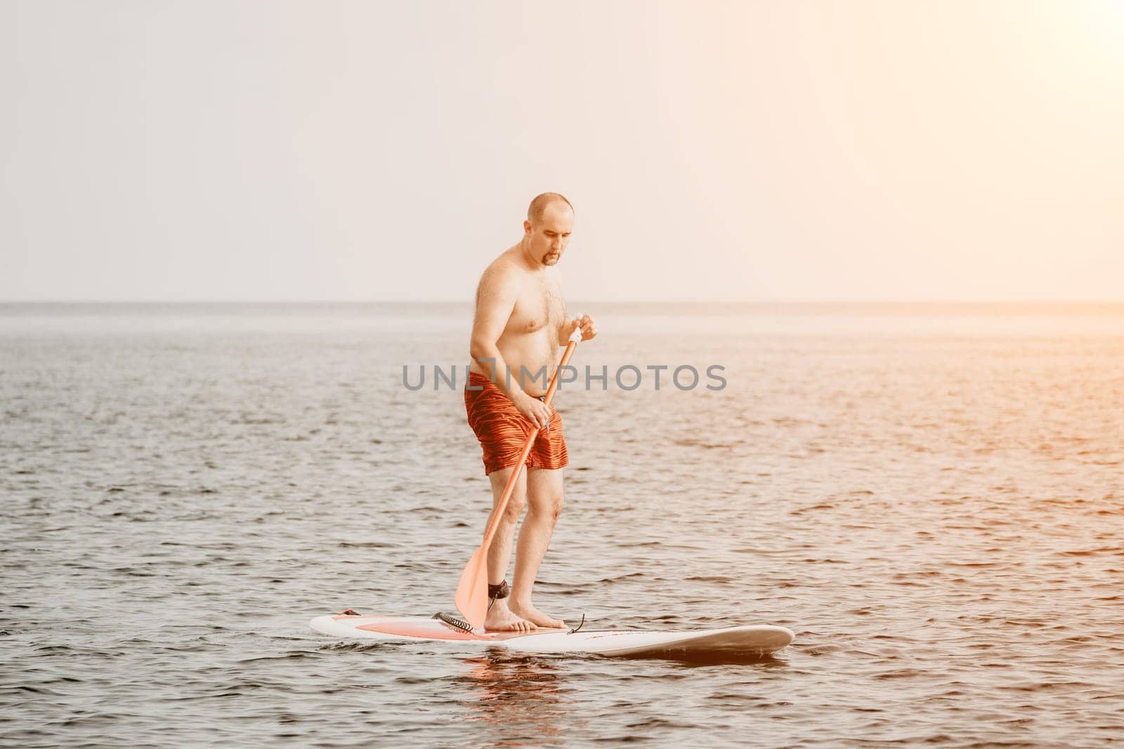 Active mature male paddler with his paddleboard and paddle on a sea at summer. Happy senior man stands with a SUP board. Stand up paddle boarding - outdor active recreation in nature