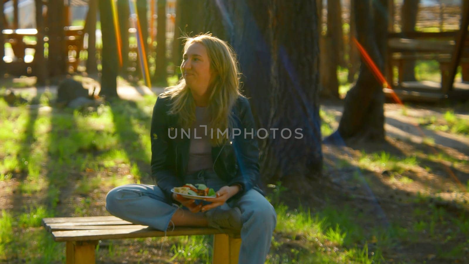 Young woman is sitting on bench and eating barbecue. Stock footage. Beautiful woman is sitting alone and eating barbecue in park in summer. Woman with barbecue plate in nature on sunny summer day.