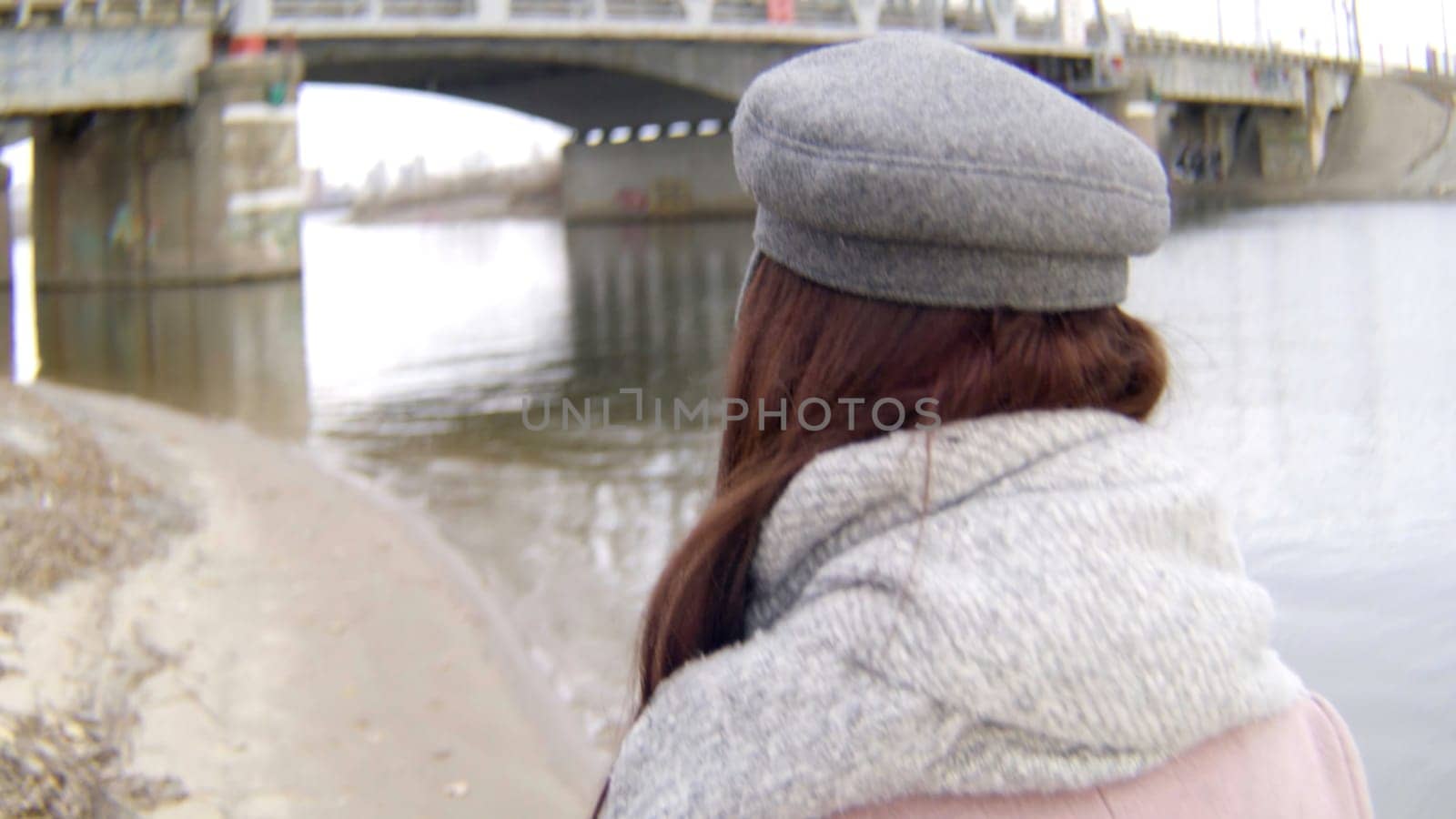 View over the shoulder of a woman in warm clothed with a yellow tree leave in hands. Stock footage. Woman walking along the beach to the bridge