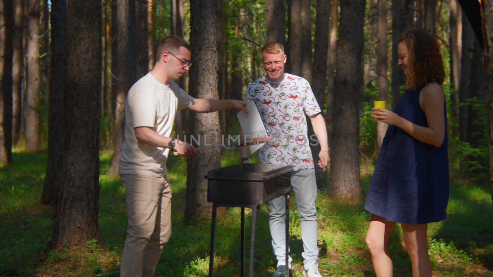 Man blows up coals on grill. Stock footage. Friends are standing at grill and blowing up coals in forest. Friends cook on grill and blow up coals for roasting meat outdoors in summer.