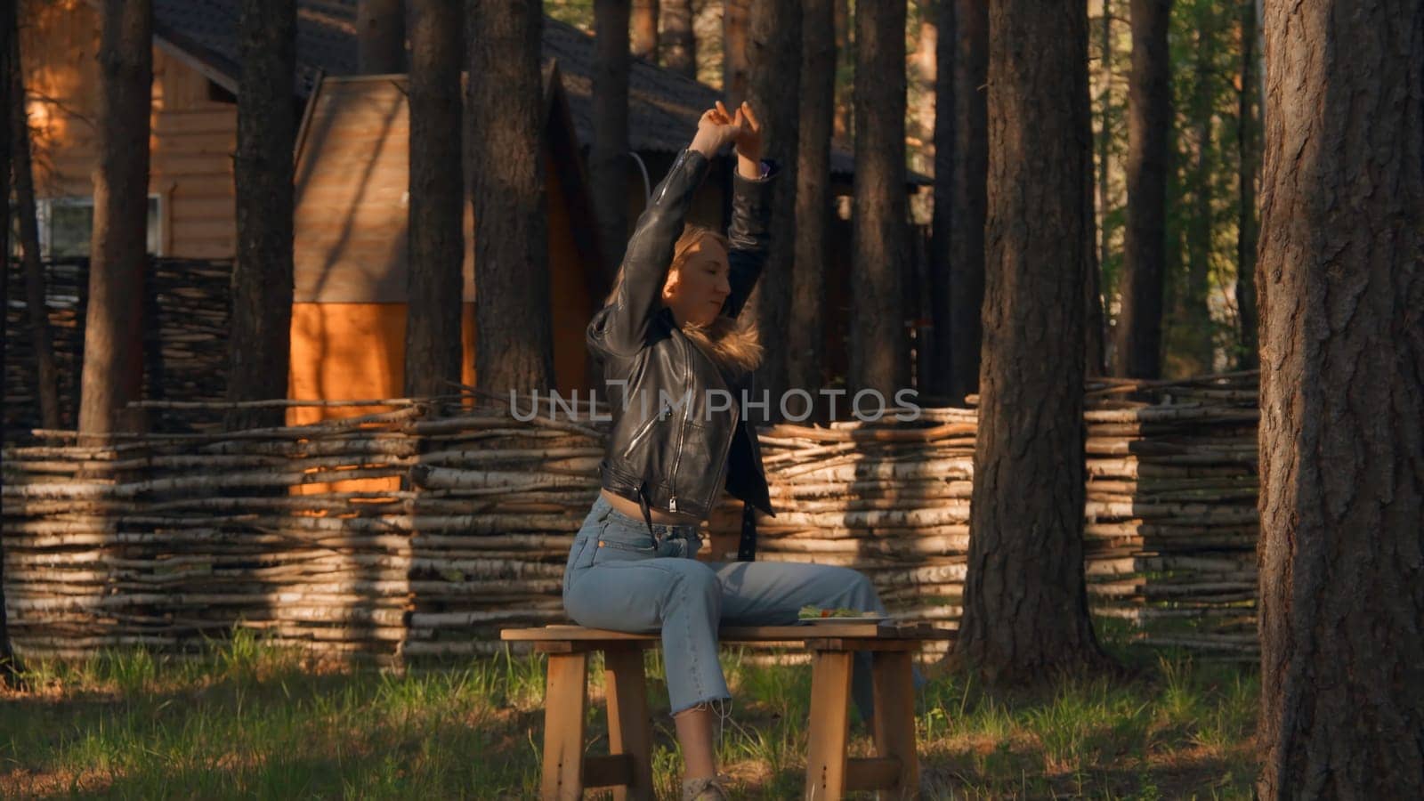 Beautiful young woman is sitting on bench and warming up. Stock footage. Young woman stretches out on bench waiting for date. Attractive woman stretches while sitting on bench in woods.
