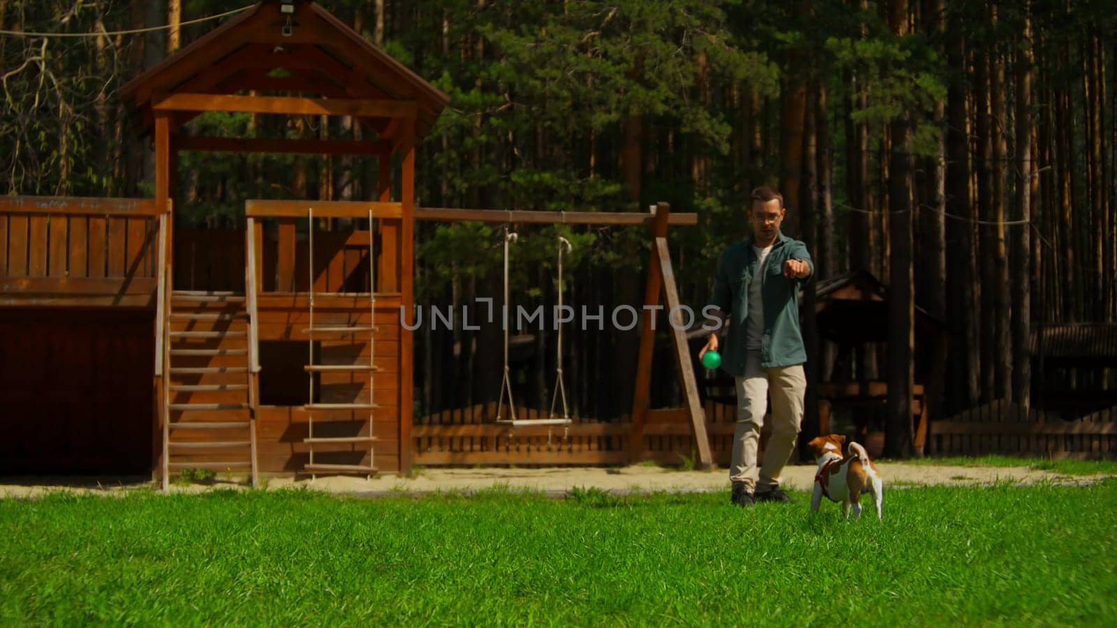 Man plays ball with dog in park. Stock footage. Man is playing ball with dog on green grass with recreation area. Outdoor activities with dog on sunny summer day.