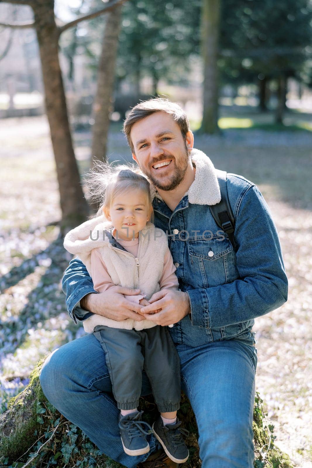 Happy dad with a little girl on his knees sit on a stump in a spring park by Nadtochiy