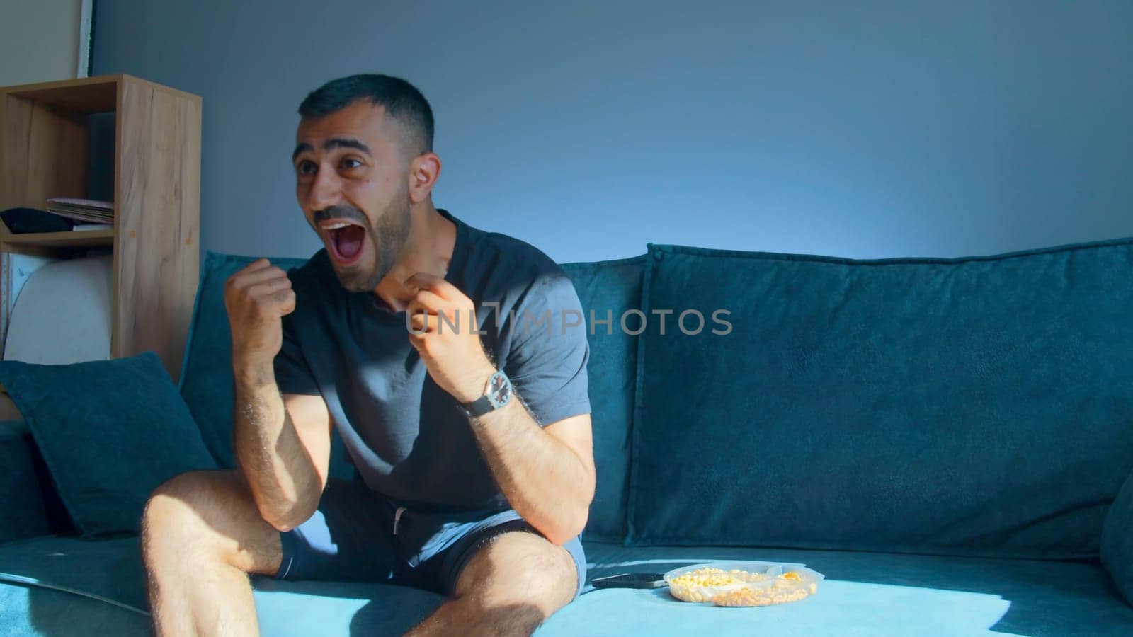 Young man enthusiastically watches football on TV. Media. Man emotionally watches football match on TV. Man eats snacks and watches football with joy.