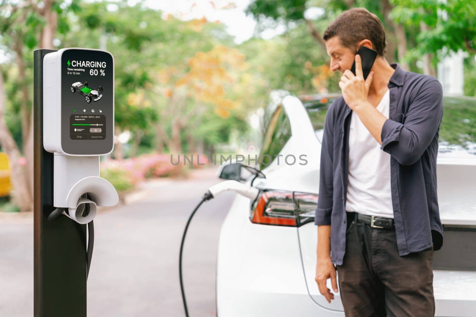 Man talking on smartphone while recharging electric car battery charging from EV charging station during vacation holiday road trip at national park or autumnal forest. Exalt