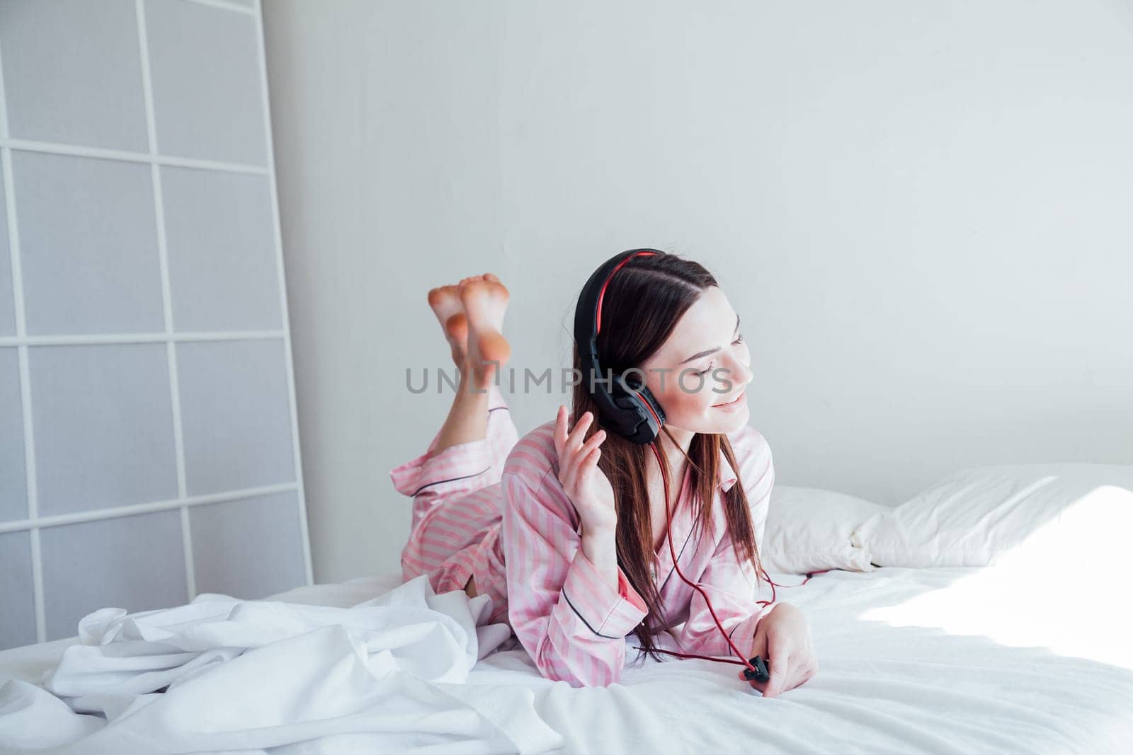 brunette woman in pink Pajamas listening to music with headphones on the bed