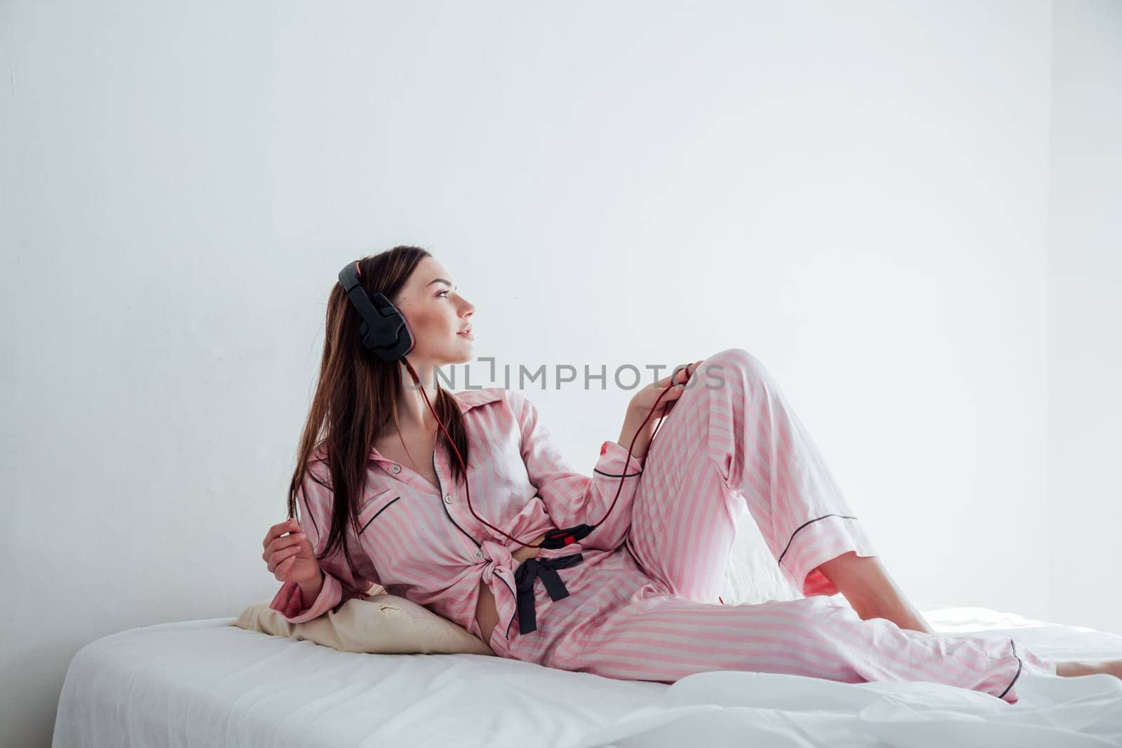 brunette in pink Pajamas listening to music in large headphones on the bed