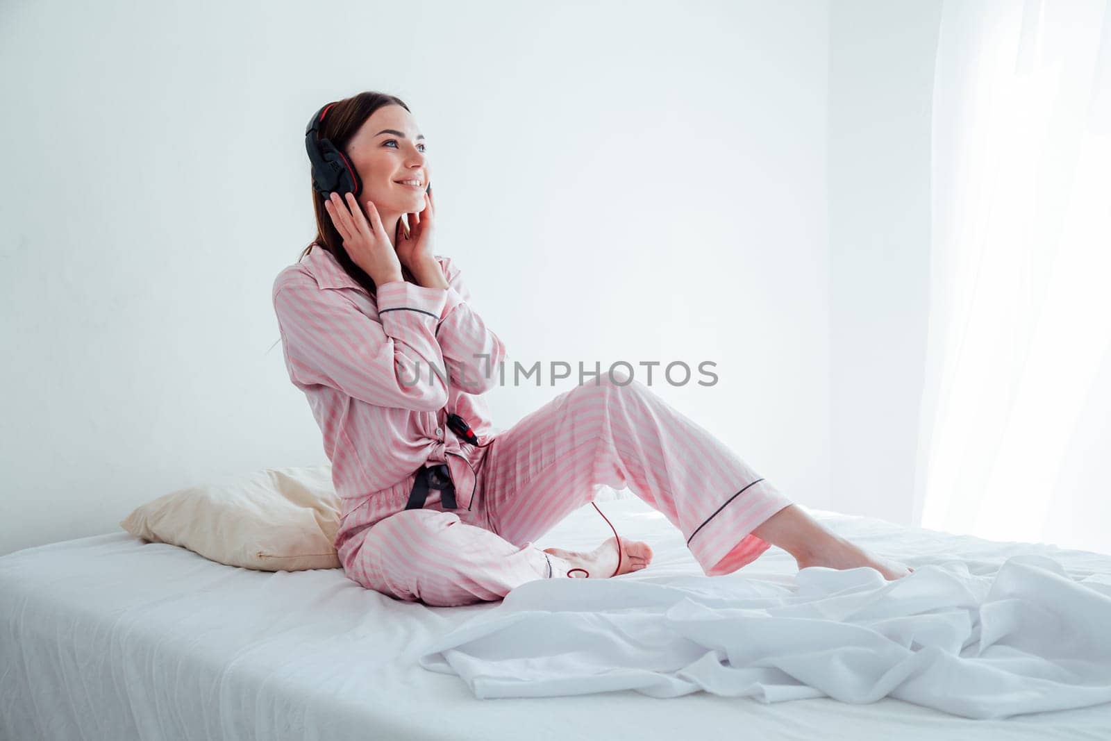 brunette in pink Pajamas listening to music in large headphones on the bed