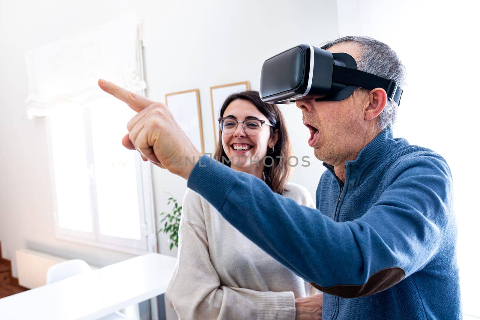 Senior man with surprise expression using virtual reality simulator at home with help of young daughter. Home and technology concept.