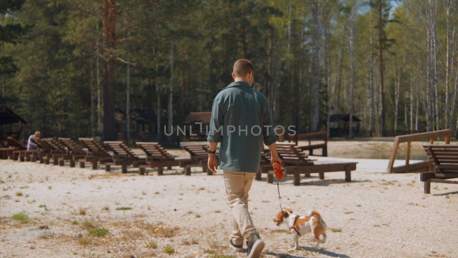 Man walks with dog on beach in woods. Stock footage. Man walks with his dog on sand at resort in summer. Man with dog walking on shore of pond in forest on sunny summer day.