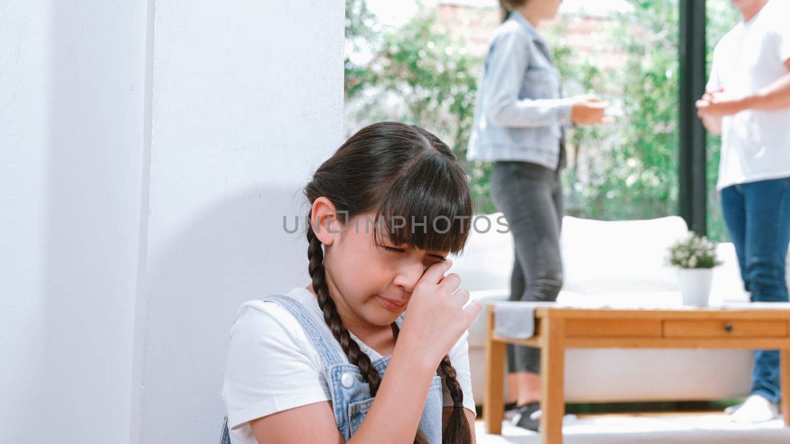 Stressed and unhappy young girl huddle in corner, cover her ears blocking sound of her parent arguing in background. Domestic violence at home and traumatic childhood develop to depression. Synchronos