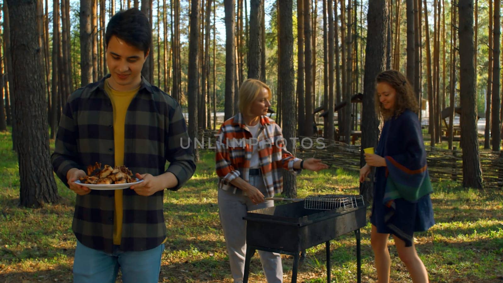 Friends cook grilled meat in forest in summer. Stock footage. Beautiful friends are relaxing in nature with barbecue in forest. Relaxing with friends in nature with barbecue on sunny summer day.