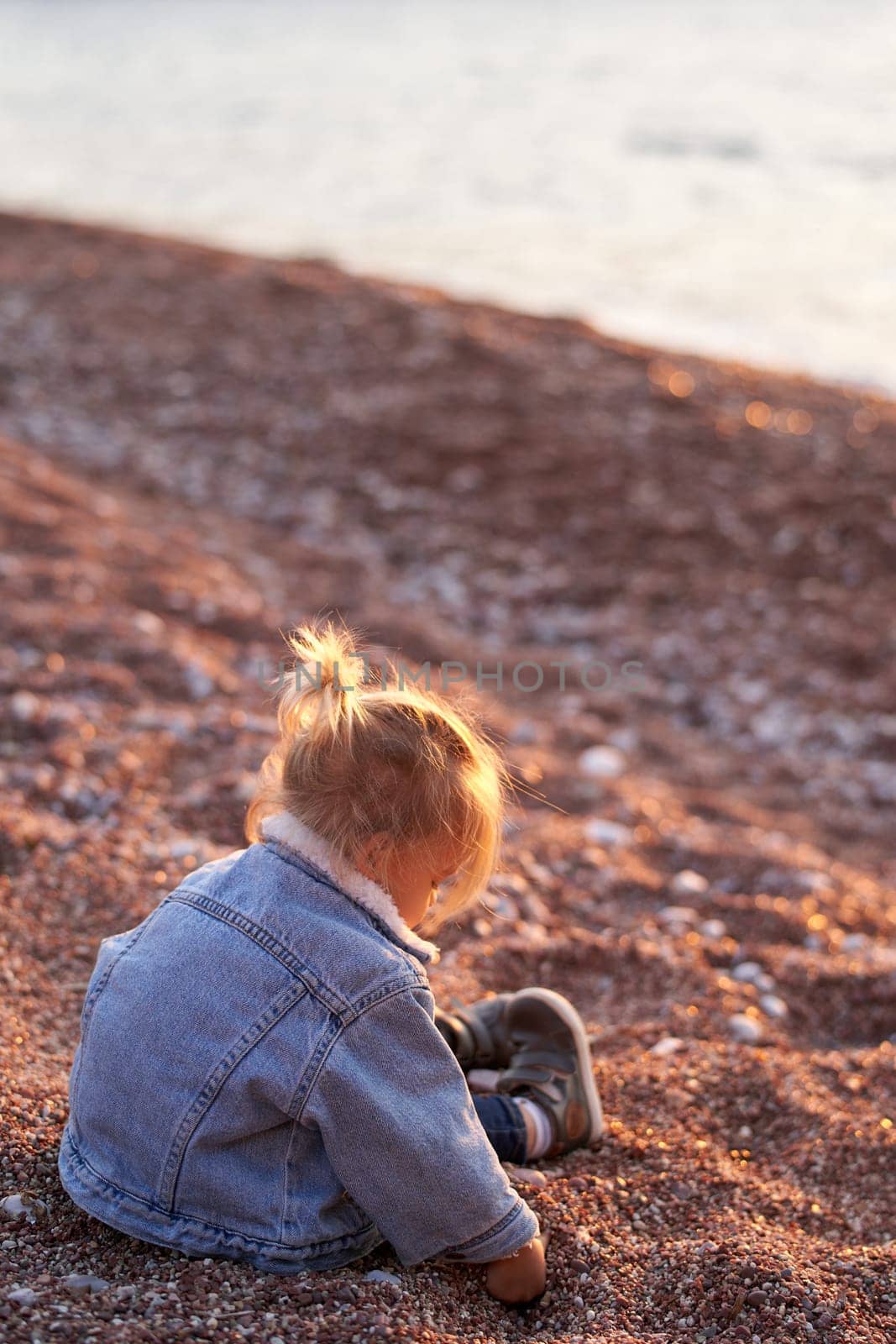 Little girl sits on the seashore and digs in small pebbles. Side view. High quality photo