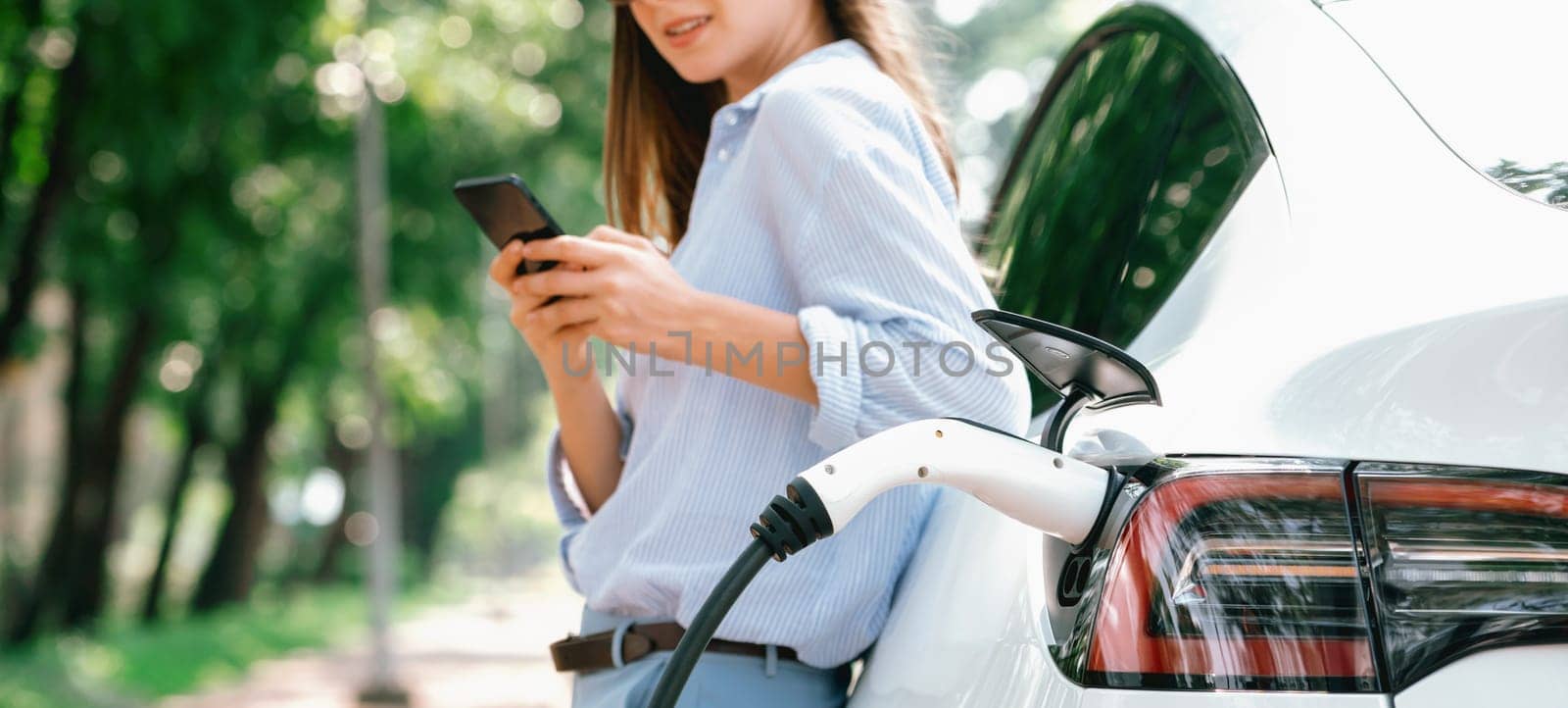 Young woman using smartphone online banking application to pay for electric car battery charging from EV charging station during vacation road trip at national park or summer forest. Panorama Exalt