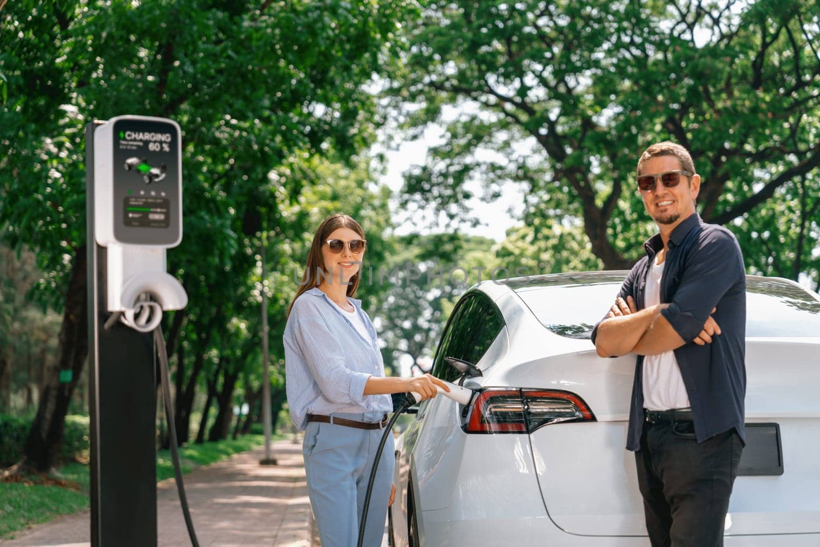 Lovely young couple wearing sun glasses recharging battery for electric car during road trip travel EV car in natural forest or national park. Eco friendly travel during vacation and holiday. Exalt