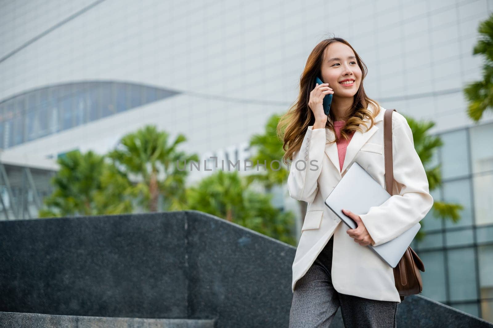 Happy successful Asian businesswoman holding laptop and Talk with cellphone on the Street next to a glass building. successfully work go to against city scene in background of modern office buildings.