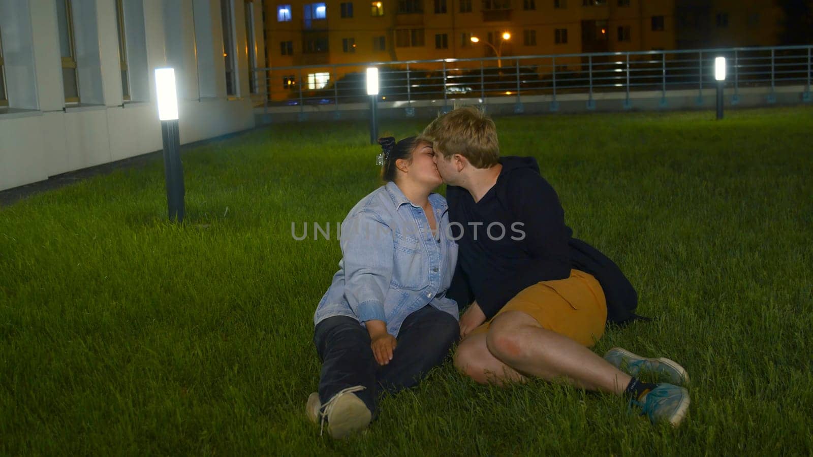 Young couple kissing on lawn. Media. Couple in love kissing sitting on green grass in park at night. Night park with kissing happy couple of overweight people by Mediawhalestock
