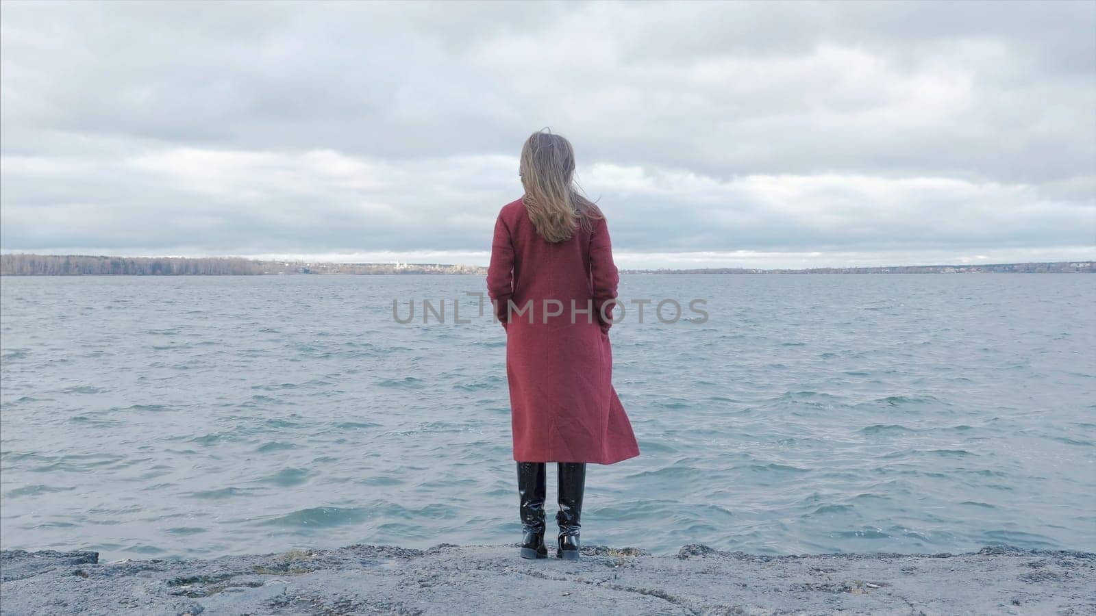 Rear view of a young woman wearing a red coat against a bright blue sky and sea on a holiday beach, outdoors. Travel and healthy lifestyle. Autumn. Girl at water, rear view p