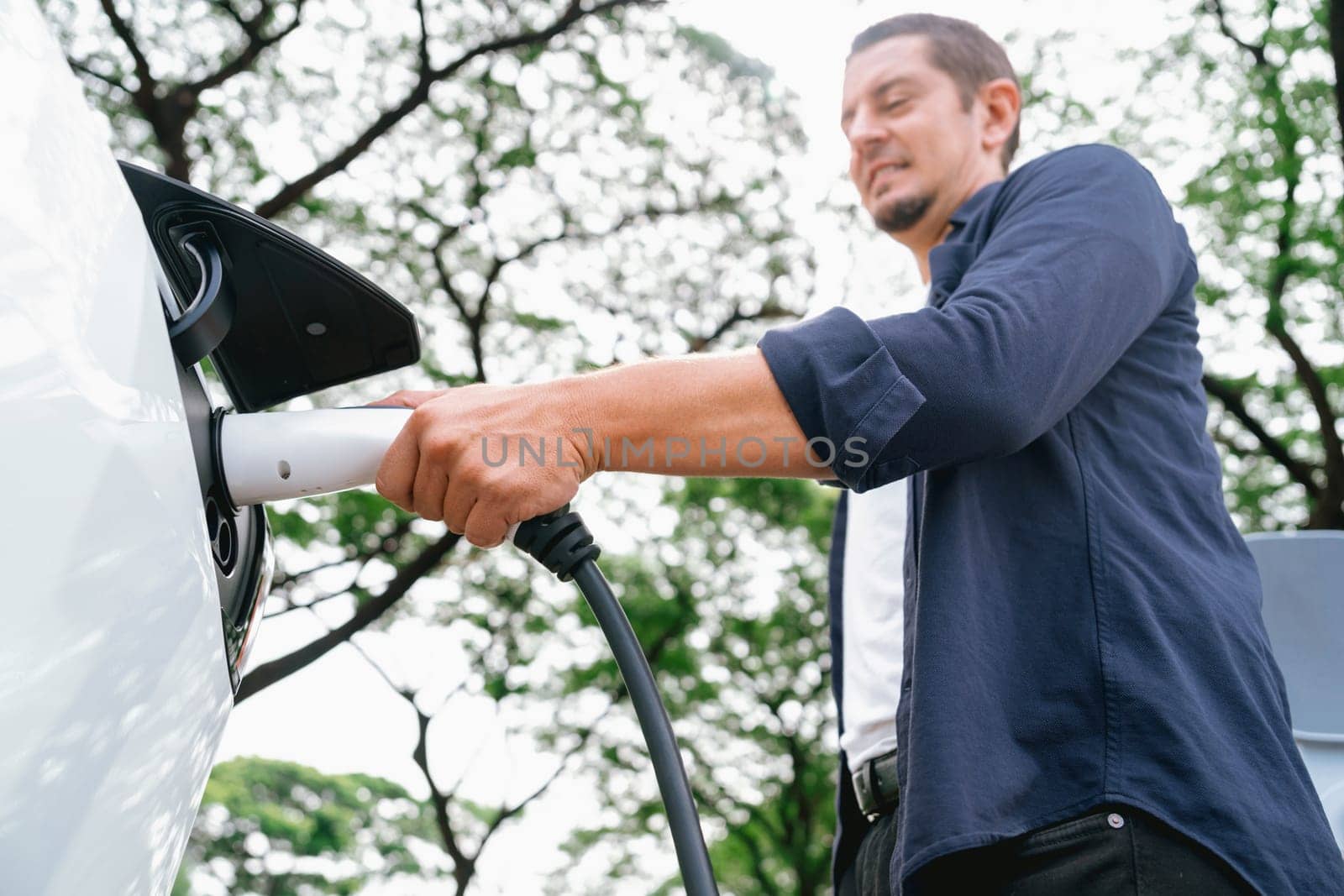 Man recharging battery for electric car during road trip travel EV car in natural forest or national park. Eco friendly travel during vacation and holiday. Exalt