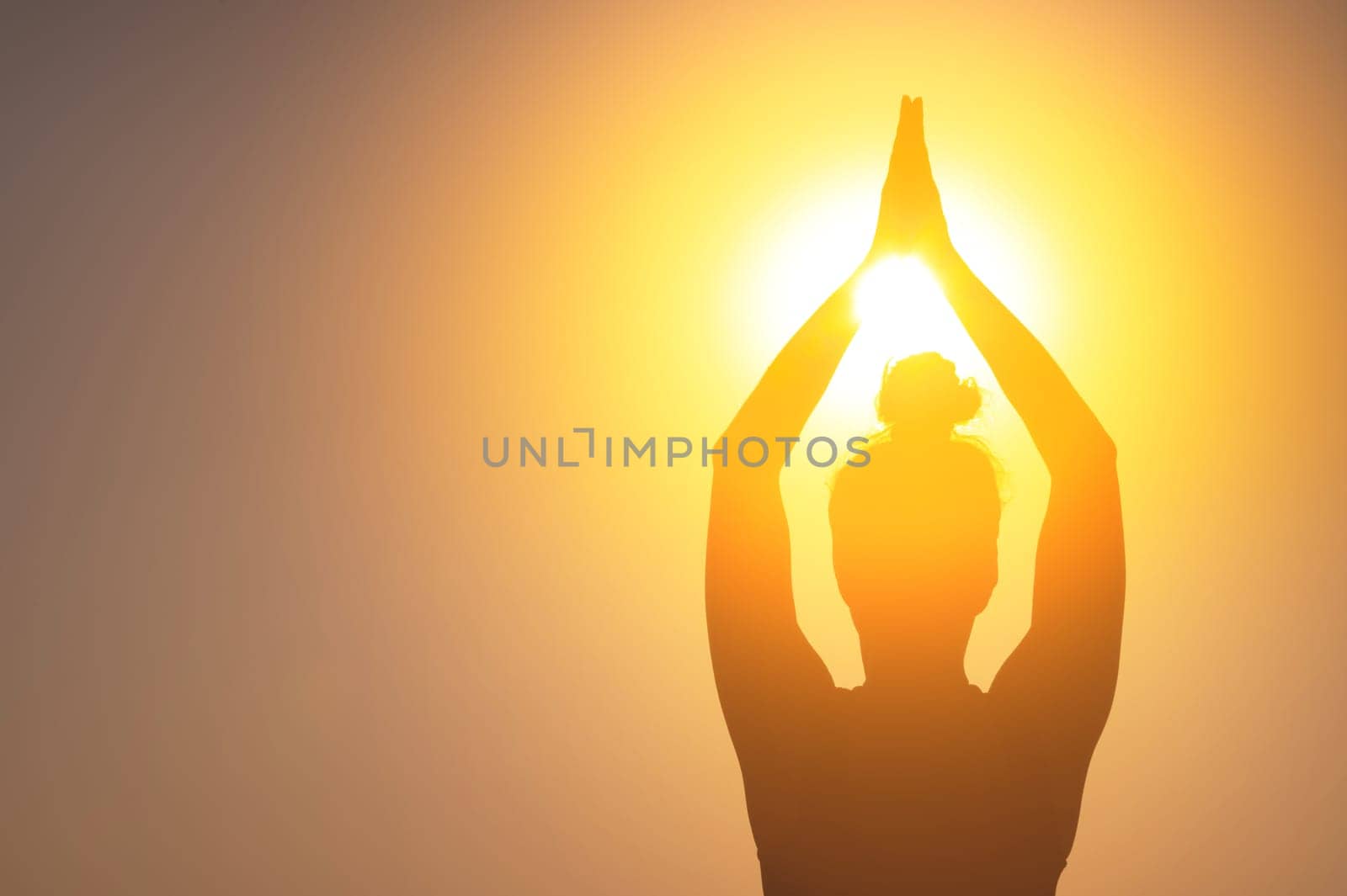 A woman meditates at sunset or dawn. Dark female silhouette with folded hands raised above her head, close-up. Light pours overhead