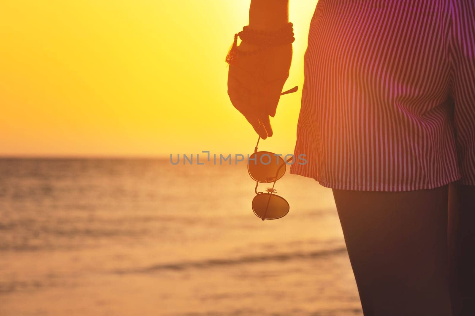 A woman in shorts stands on the shore with sunglasses in her hand, close-up. Warm colors, beach holiday at sunset.