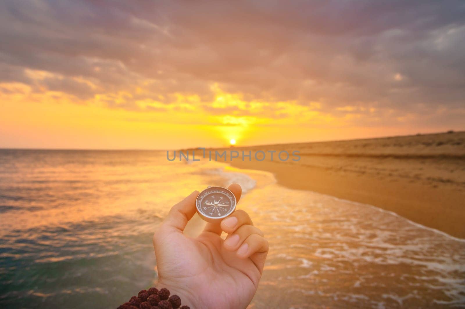 hand holds a compass against the background of the sea. the setting sun falls on the beach and ocean, there is no one on the shore by yanik88