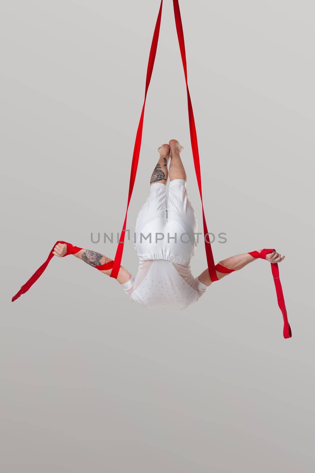 Sportsman performance on a red canvases. Tattoed man in a light sport suit is doing an acrobatic elements hanging on a rope in a studio isolated on white background. Dancing in the air with balance.