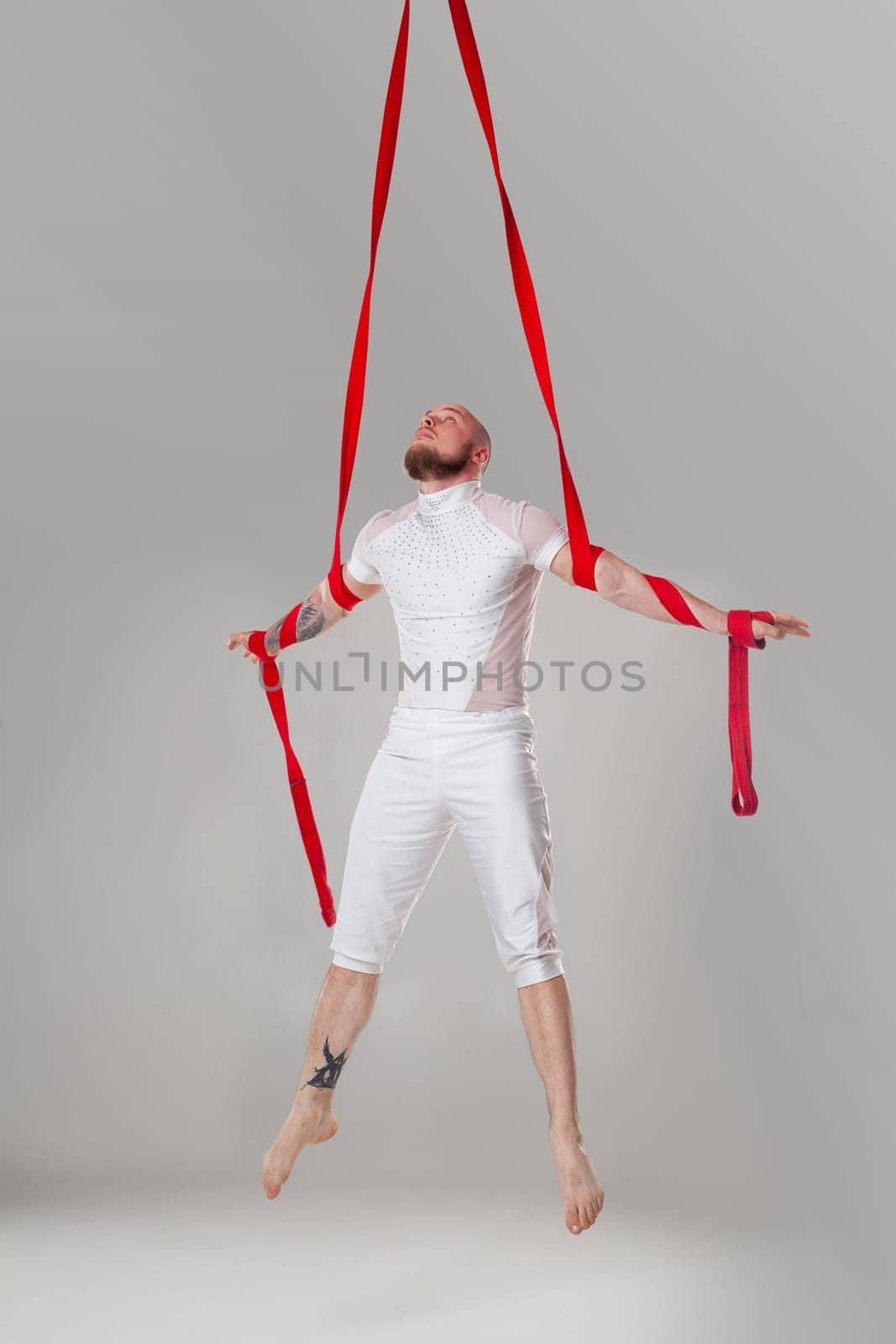 Gymnast performance on a red canvases. Strong man in a light sport suit is doing an acrobatic elements hanging on a rope in a studio isolated on white background. Dancing in the air with balance.
