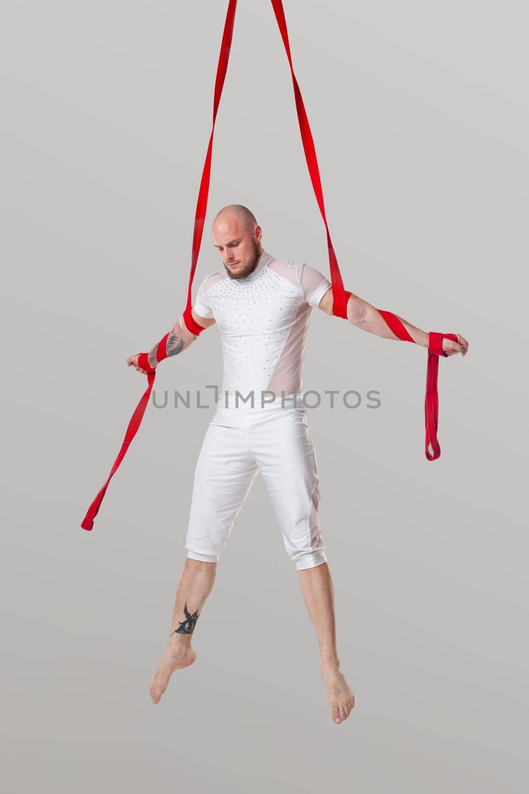 Sportsman performance on a red canvases. Bald male in a light sport suit is doing an acrobatic elements hanging on a rope and looking down in a studio isolated on white background. Dancing in the air with balance.