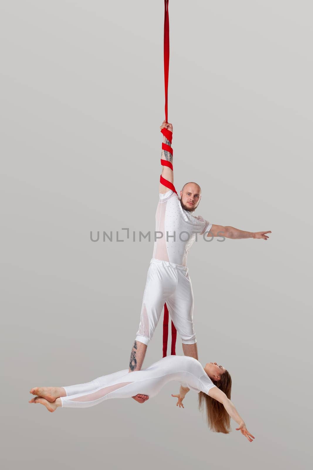 Gymnasts performance on a long red canvases. Attractive girl and an athletic man in a light sport suits are doing acrobatic elements in a studio isolated on white background. He is holding her by his legs. Dancing in the air with love and balance.